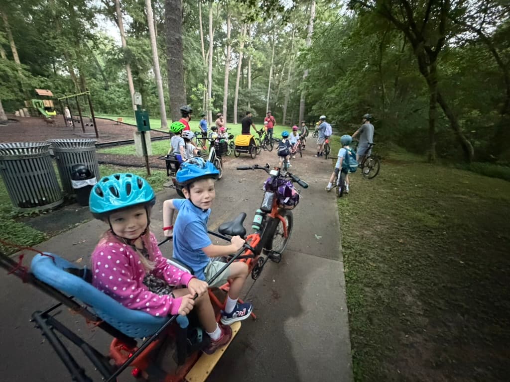 Two young riders in a cargo bike seat at the park meetup, with the group gathering behind them