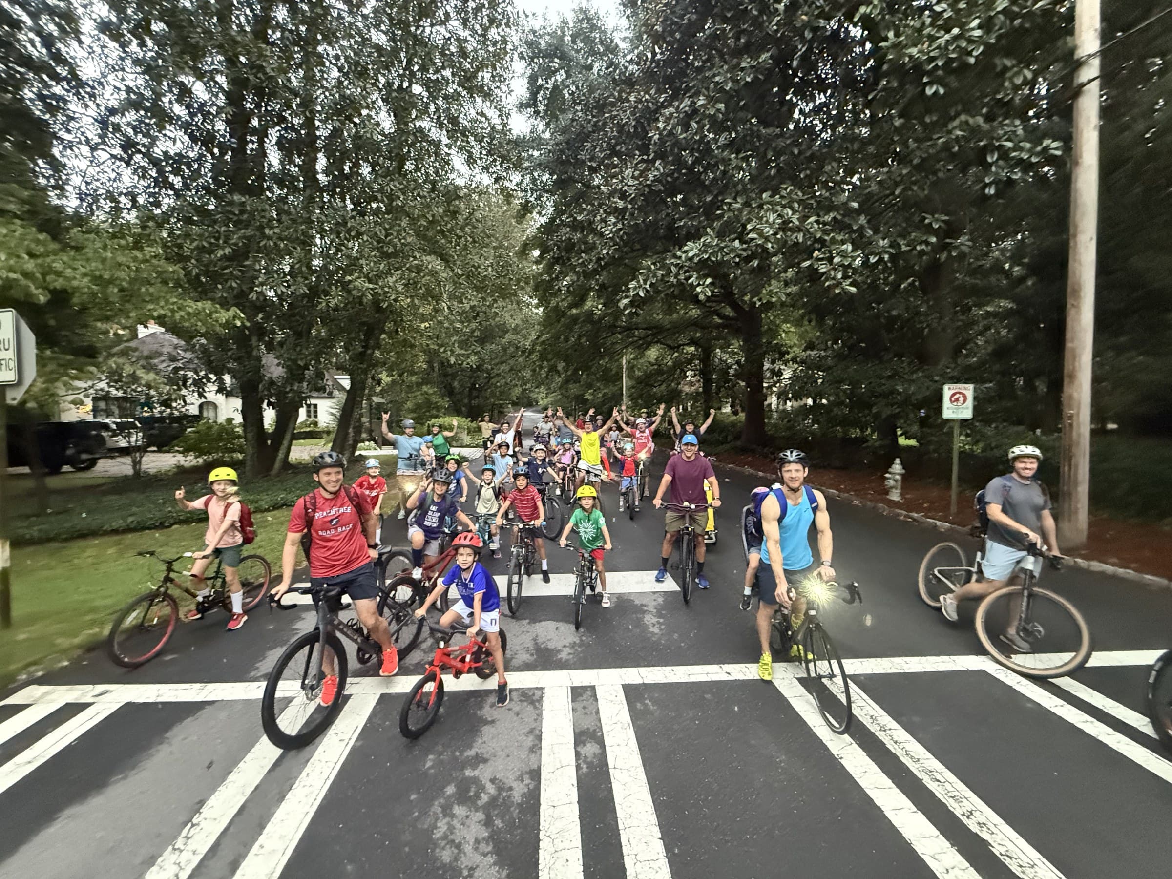 The whole bike bus stopped at a crosswalk on a tree-lined street, kids and parents cheering with their arms in the air